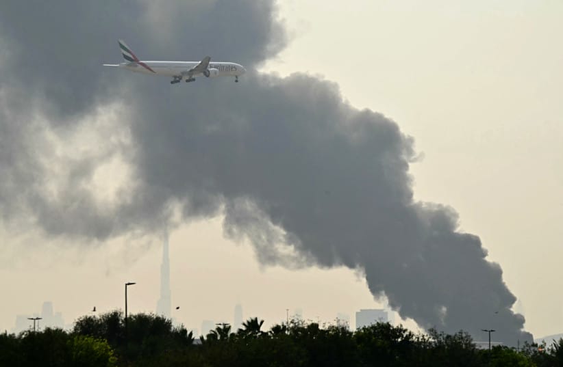An Emirates aircraft flies past plumes of smoke from an ongoing fire near Dubai International Airport in Dubai due to an Iranian attack on March 16, 2026.