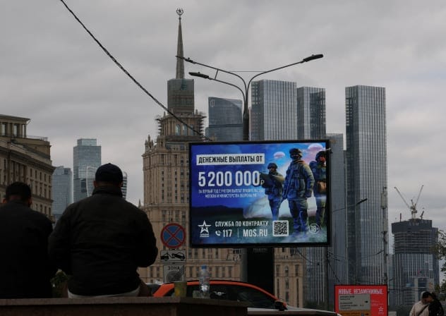 Men sit in front of an advertising board promoting military service under contract in the Russian Armed Forces and informing about payments, against the skyline of downtown Moscow, Russia, August 27, 2026.