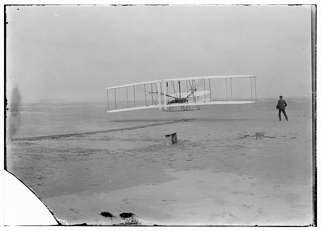 Black and white photograph shows plane several feet off the ground.  A person is standing in the background watching.