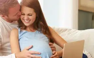 זוג בהריון יושבים ביחד על הספה עם לפטופ ומחייכים