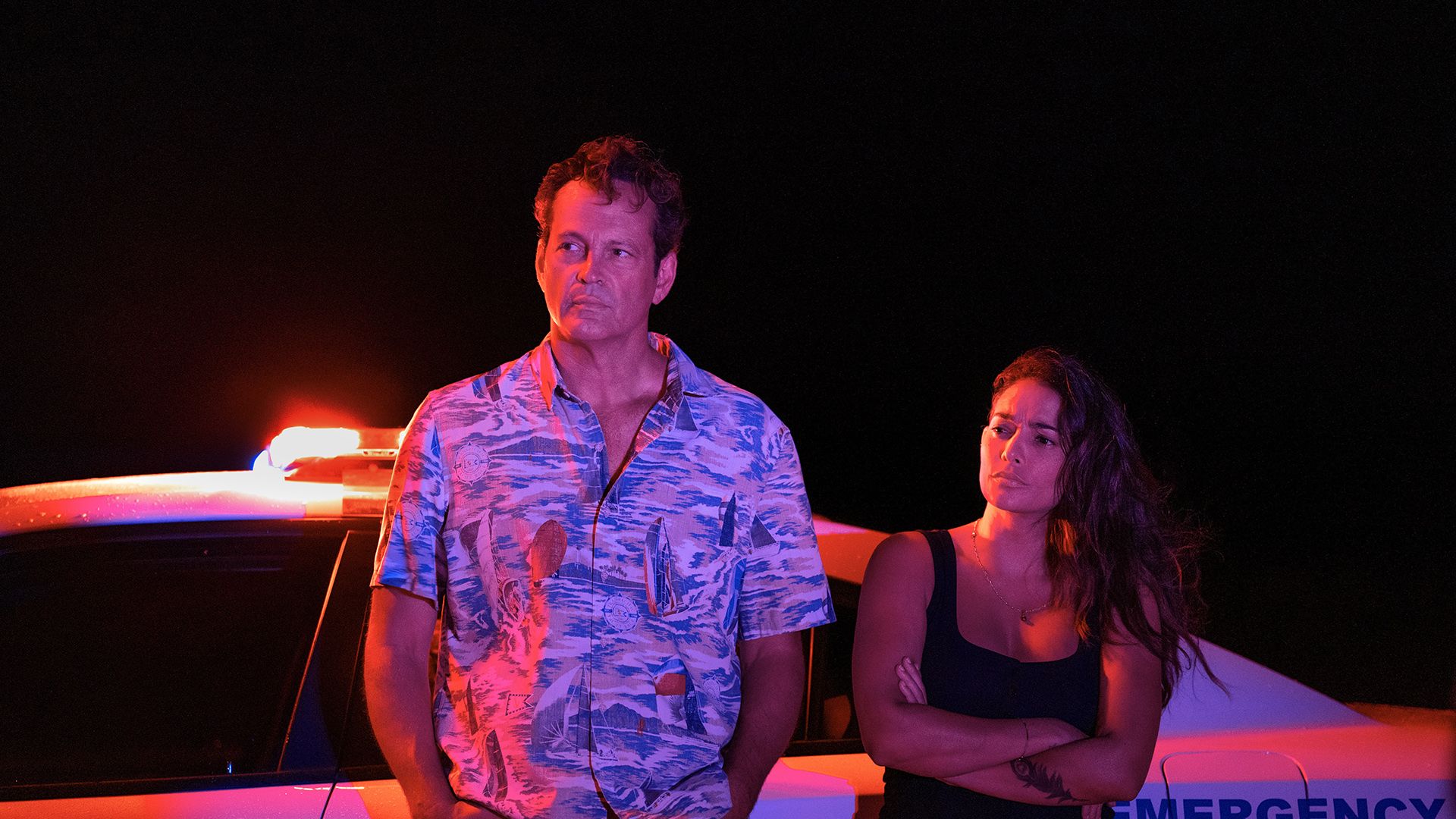 A dark image of Andrew and Rosa from Bad Monkey outside at night, leaning against a police car.