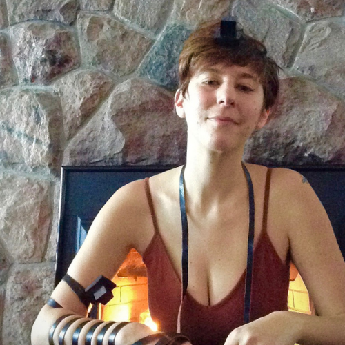 Sarit sits in front of a fireplace with a stone wall around it. They are wearing tefilin on their arm and head and a red tank top.