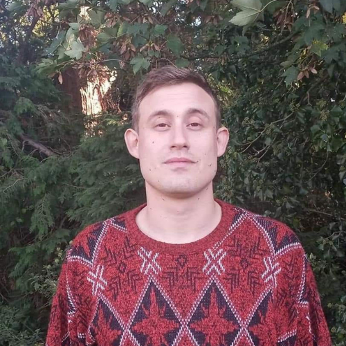Lev stands in front of a green, leafy background. He is wearing a red pattered sweater.