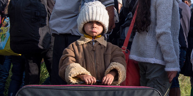 Ein kleiner Junge mit riesiger weißer Mütze steht hinter einem großen Koffer, hinter ihm widerum stehen Erwachsene, die ihm den Rücken zukehren Ein kleiner Junge mit riesiger weißer Mütze steht hinter einem großen Koffer, hinter ihm widerum stehen Erwachsene, die ihm den Rücken zukehren